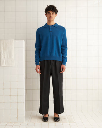 A person stands in a tiled room, wrapped in a cozy Cashmere Polo - Ocean and black pants, with a towel hanging nearby.