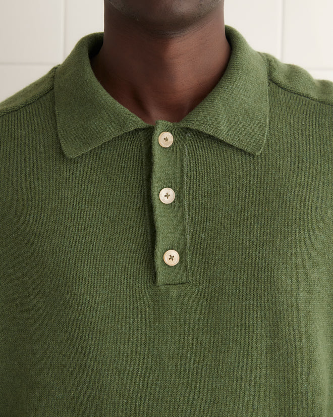 A person wearing a green Cashmere Polo and blue jeans stands on a tiled floor, with a towel hanging on the white tiled wall beside them.