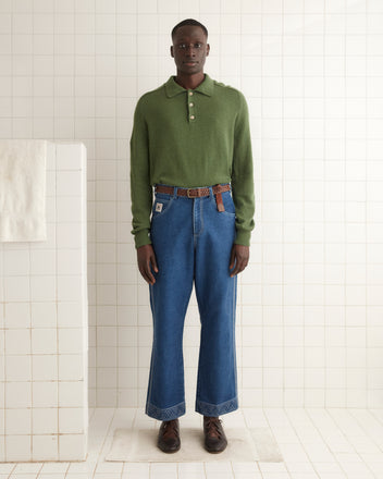 A person wearing a green Cashmere Polo and blue jeans stands on a tiled floor, with a towel hanging on the white tiled wall beside them.