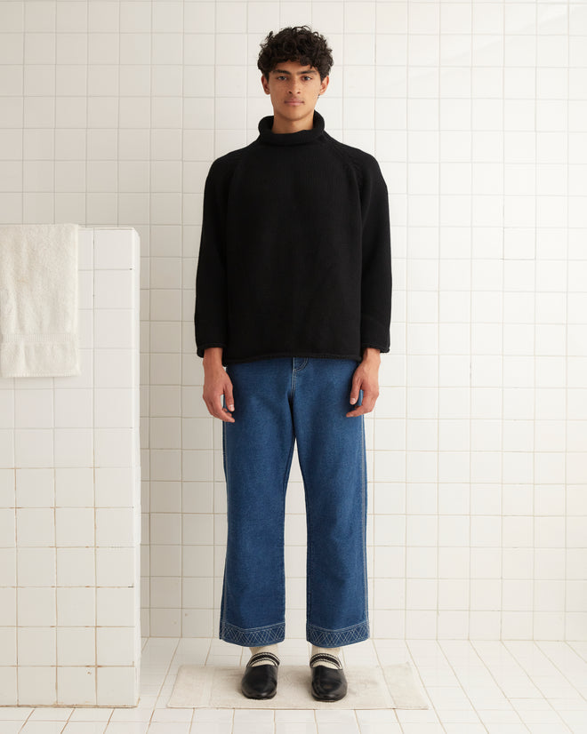 A person wearing the Cashmere Rollneck - Black sweater and jeans stands in a tiled room with a towel on a rack.