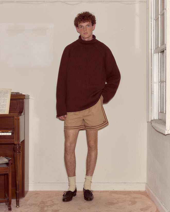 Someone in an oversized fit Cashmere Rollneck - Dark Brown sweater and tan shorts stands indoors beside a piano and a window.