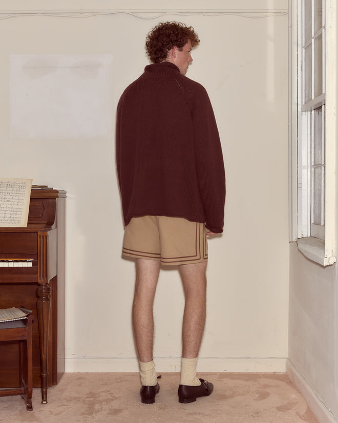 A person stands indoors facing a wall, wearing an oversized fit Cashmere Rollneck - Dark Brown sweater, beige shorts, socks, and dark shoes.