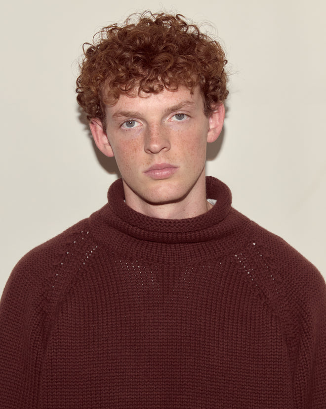 A young man with curly red hair wears the Cashmere Rollneck - Dark Brown, styled in an oversized fit, looking seriously at the camera against a plain background.