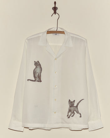 The Cat Motif Long Sleeve Shirt is a white, vintage-inspired button-up featuring illustrations of two playful gray cats, displayed on a hook against a plain wall.