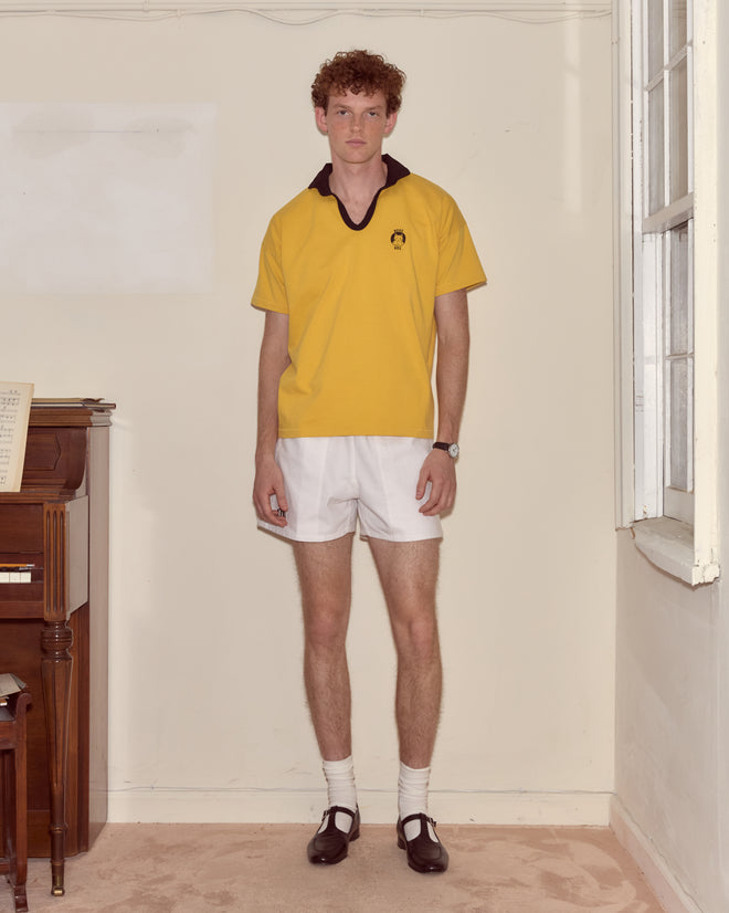 A young man stands indoors by a piano wearing the Cat Polo - Yellow, made from 100% cotton, paired with white shorts, socks, and black shoes.