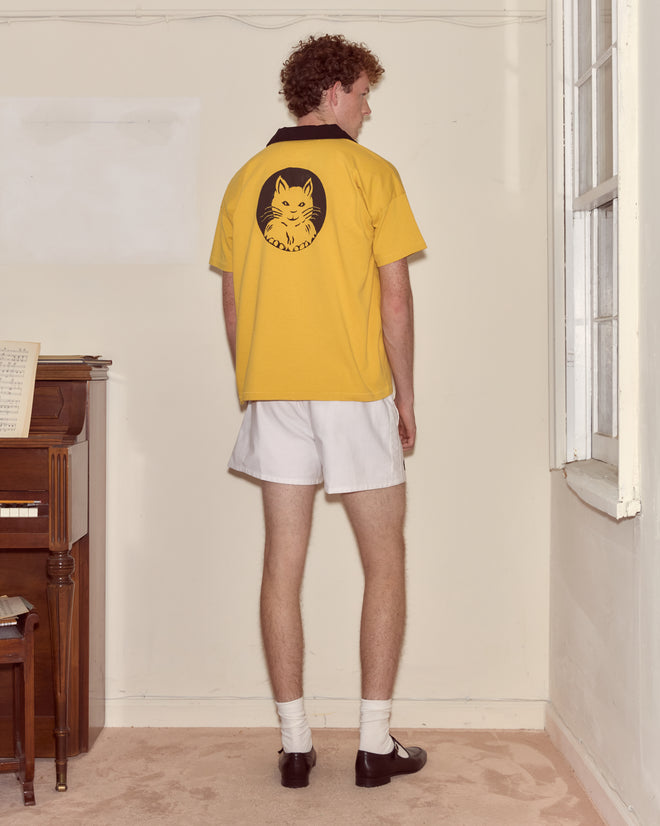 A person in a yellow Cat Polo and white shorts stands facing a wall near a piano and window.