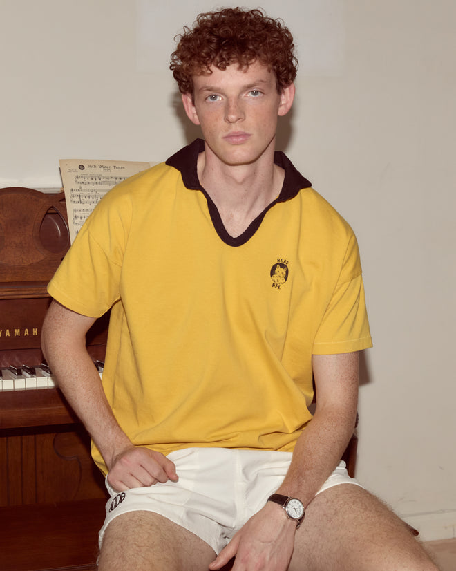 A young man with curly hair, wearing a Cat Polo - Yellow and white shorts, sits by a piano with sheet music on it.