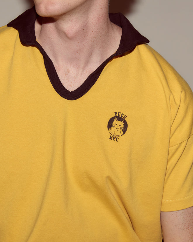 A person wears the Cat Polo - Yellow: a 100% cotton yellow shirt with a black collar, featuring a Cheshire Cat polo logo on the left chest and "BODE REC" text.