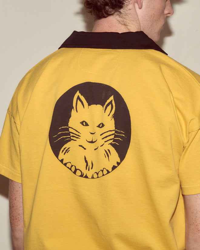 Someone wears the Cat Polo - Yellow, featuring a black collar and a cartoon cat face graphic on the back, styled in a rugby silhouette.