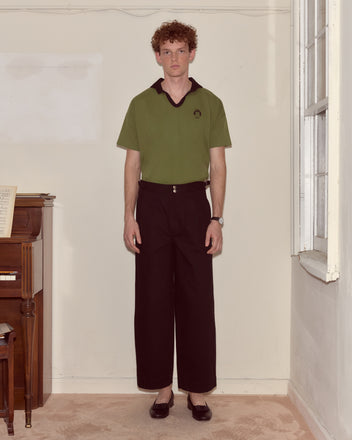 A person with curly hair stands indoors in a Cat Polo - Green and black pants, near a piano and a window.