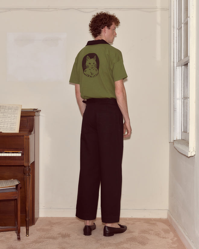 A man stands indoors facing away, wearing a Cat Polo - Green with black pants.