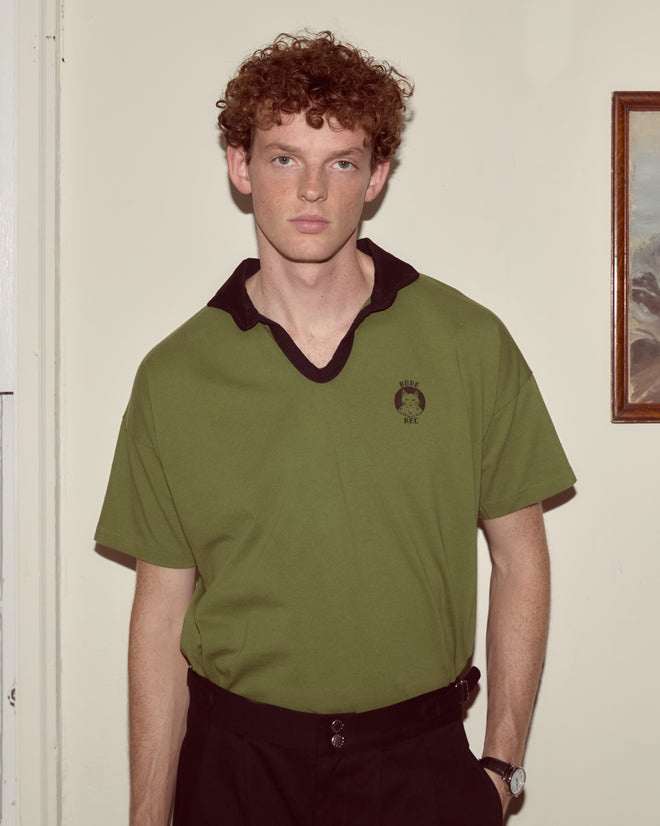 A young man with curly red hair stands indoors by a wall and paints while wearing the Cat Polo - Green shirt and black pants.