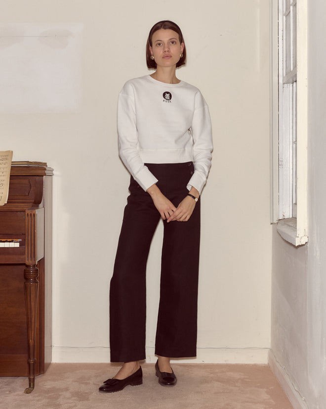 A woman stands indoors near a piano, wearing the Cat Pullover made from heavyweight fleece and black pants, with her hands clasped in front.