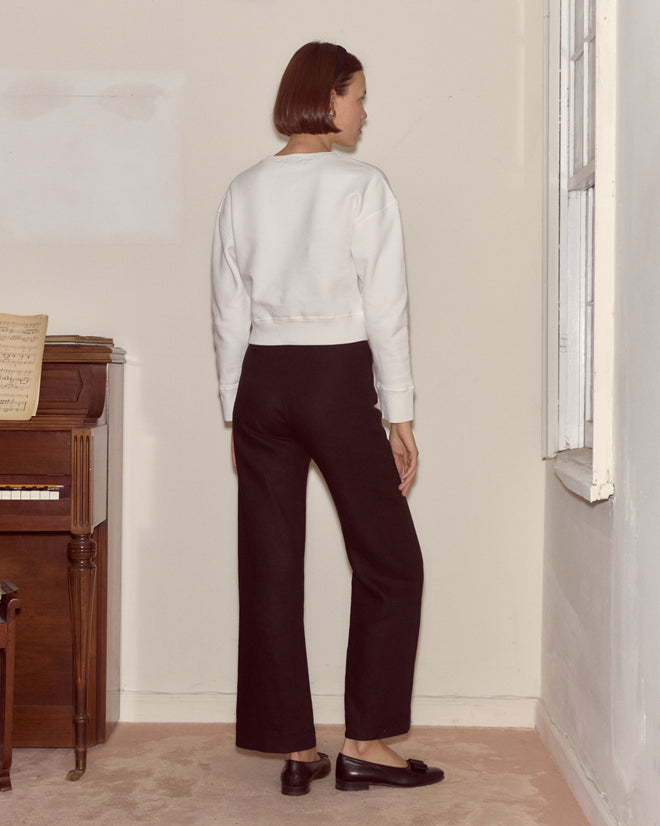 A woman stands indoors facing a wall, wearing a white Cat Pullover heavyweight fleece sweater, black pants, and black shoes.