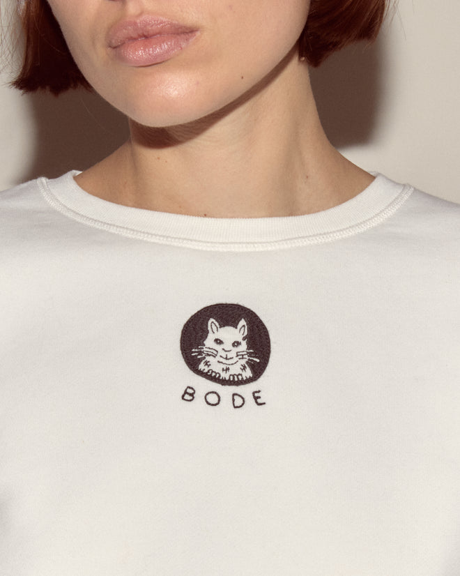 A woman in a heavyweight Cat Pullover, featuring a white shirt design with "BODE" and a charming embroidered patch on the front.
