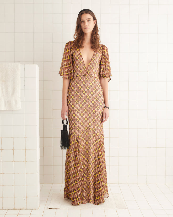 A woman in a 1930s-inspired Cattail Plaid Fair Gown with a deep V-neck holds a small black bag, standing against a tiled wall.