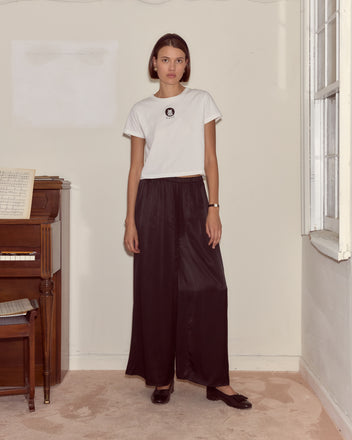 A woman in a Cat Tee and black wide-leg pants stands by a piano in a softly lit, minimal room.