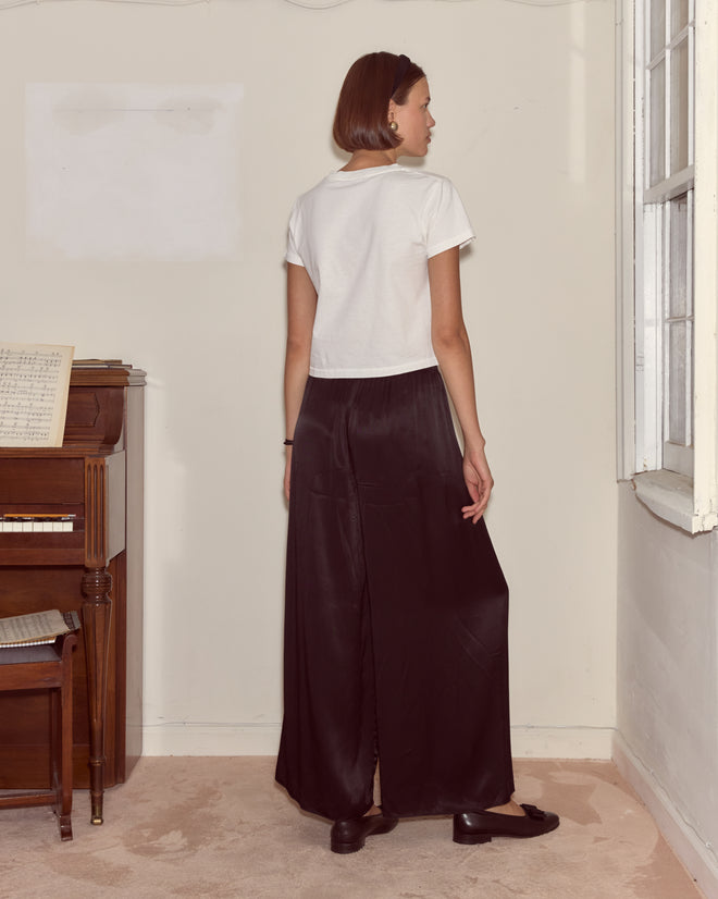 A woman with short hair, wearing a fitted Cat Tee and black skirt, stands indoors facing away from the camera.
