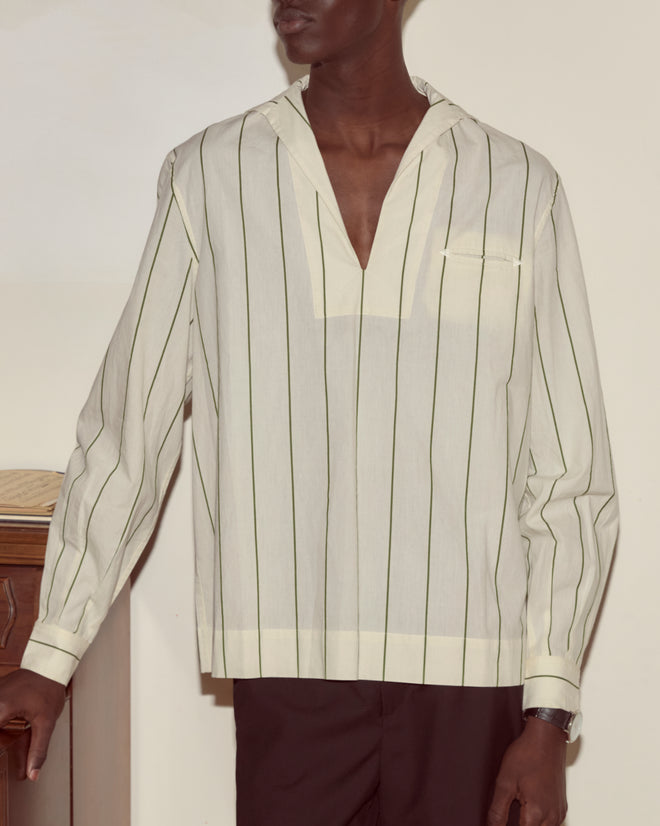 A person in the Celery Stripe Cricket Shirt—a cream, long-sleeve, 100% cotton V-neck with stripes—and dark trousers stands by a wooden table.
