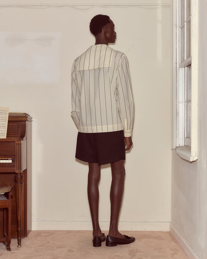 Wearing the Celery Stripe Cricket Shirt and black shorts, a person stands facing the corner of a beige room near a piano and window.
