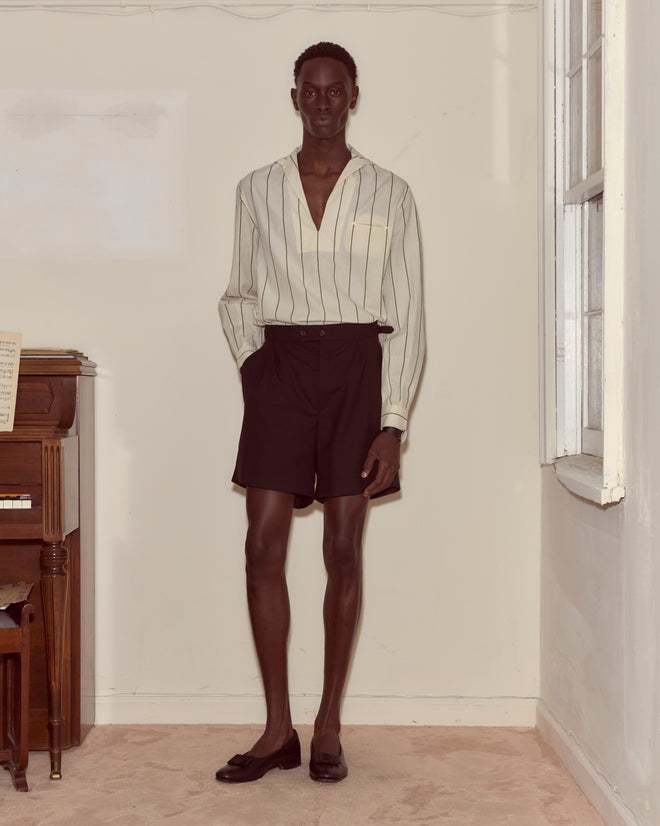 A person stands indoors by a piano wearing the Celery Stripe Cricket Shirt, black shorts, and loafers.