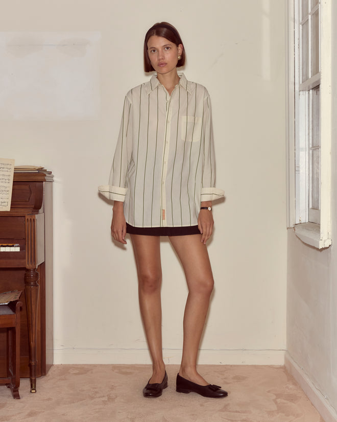 A woman wearing the Celery Stripe Murray Shirt and a black skirt stands indoors beside a piano, near a window, on a beige carpet.