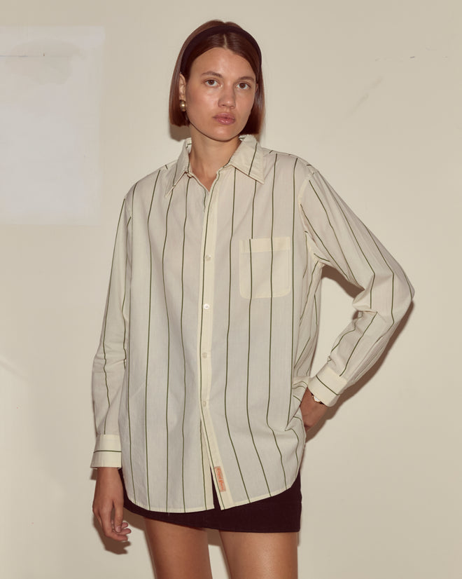 Woman wearing the Celery Stripe Murray Shirt with an antique City of Paris label and a black skirt, standing against a plain beige wall.