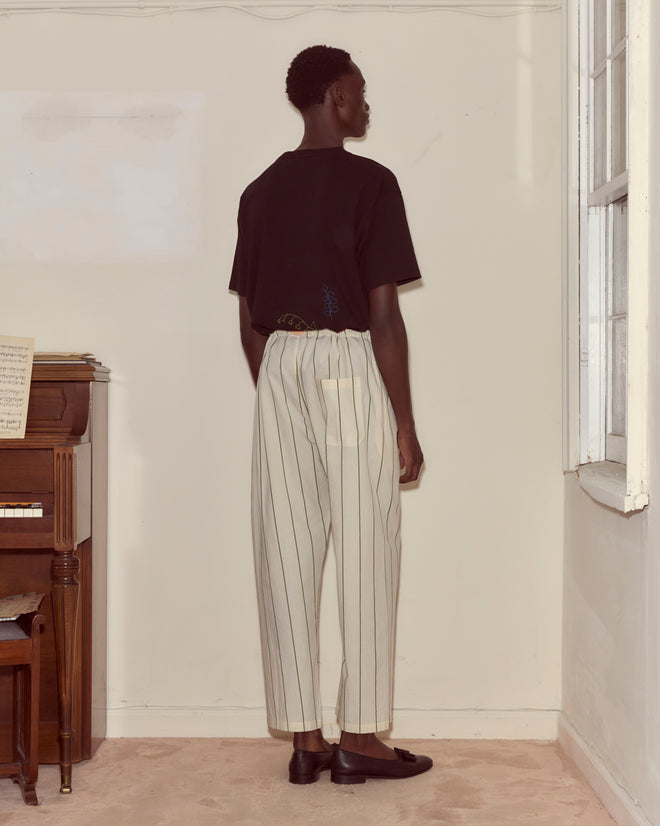 A person stands indoors near a window and a piano, facing away, wearing a black shirt and Celery Stripe Pajama Pants made of 100% cotton.