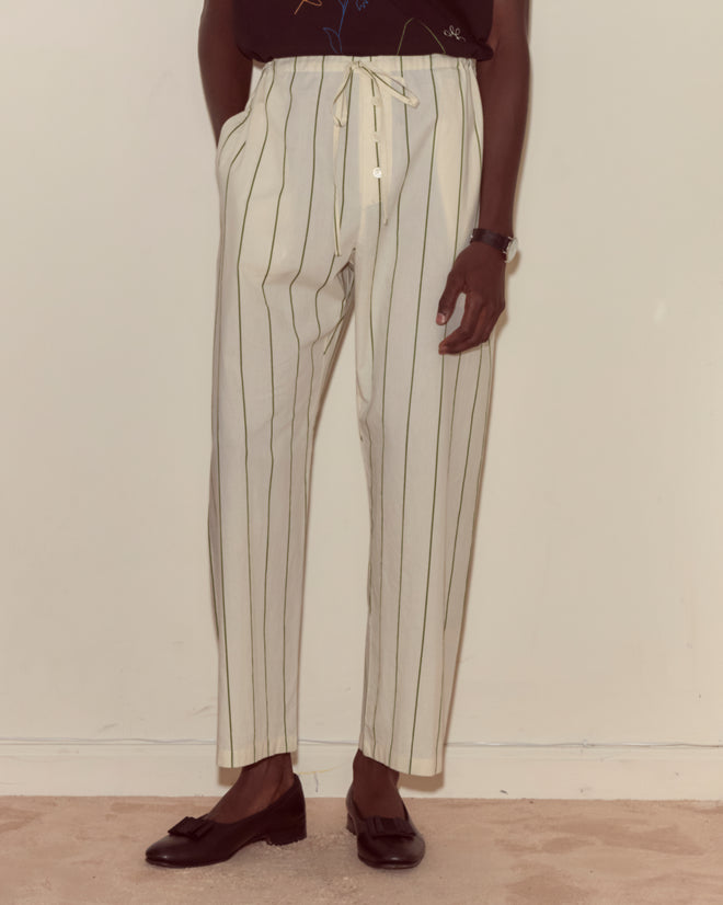 Someone wearing Celery Stripe Pajama Pants—cream-colored with vertical stripes, made from 100% cotton—paired with black loafers, stands against a plain wall.