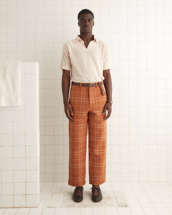 A man stands in a tiled room wearing an off-white short-sleeve shirt, Centennial Plaid Trousers made from 1970s suiting fabric, a brown belt, and brown shoes.