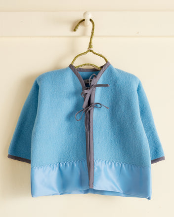 A Cerulean Split Baby Jacket - OS with grey trimmings and a tie closure, crafted from fuzzy wool, hanging on a white hook.