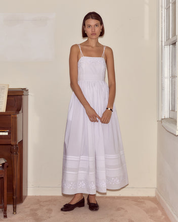 A woman in the Chamomile Dress, made of white cotton poplin, stands indoors by a piano and window, looking at the camera.