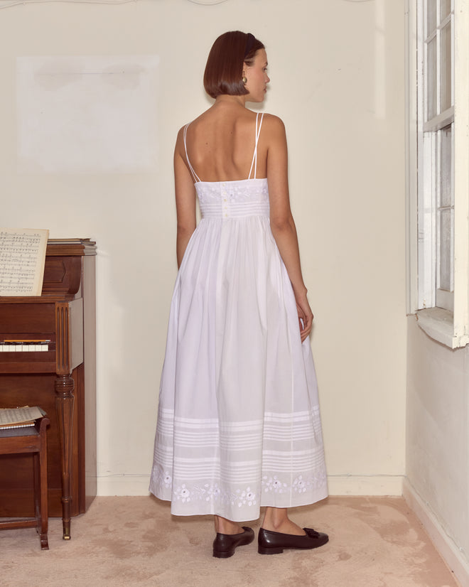 A woman stands indoors near a piano and window, facing away, wearing the Chamomile Dress—a white, sleeveless cotton poplin style.