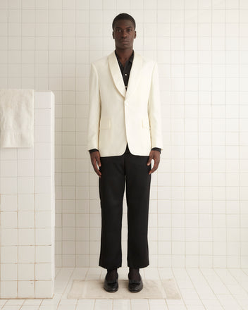 Wearing the Chantilly Tuxedo Jacket, a person poses elegantly against a pale wall in a softly lit room, completing their look with a white shawl-collar jacket, red vest, and fine wool loafers.