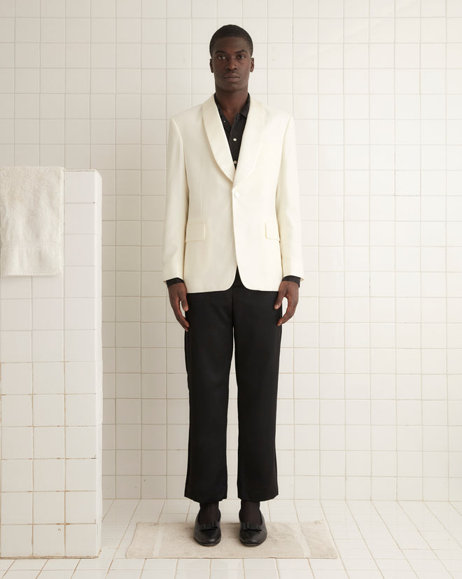 Wearing the Chantilly Tuxedo Jacket, a person poses elegantly against a pale wall in a softly lit room, completing their look with a white shawl-collar jacket, red vest, and fine wool loafers.