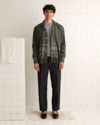 A person in a charcoal fringe long sleeve shirt and dark trousers stands in a tiled room, their gaze wandering over an antique carriage blanket draped alongside a towel on the wall partition.