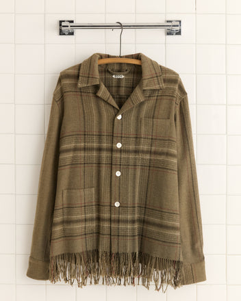 A Charcoal Fringe Overshirt - S/M, made of wool flannel with a fringe hem, hangs on a hanger against a tiled wall, reminiscent of an antique carriage blanket's charm.