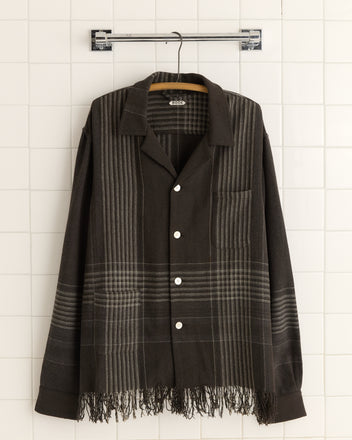 A unique Charcoal Plaid Overshirt in XL/XXL, featuring a fringe hem reminiscent of an antique carriage blanket, elegantly hangs on a hanger against a tiled wall.