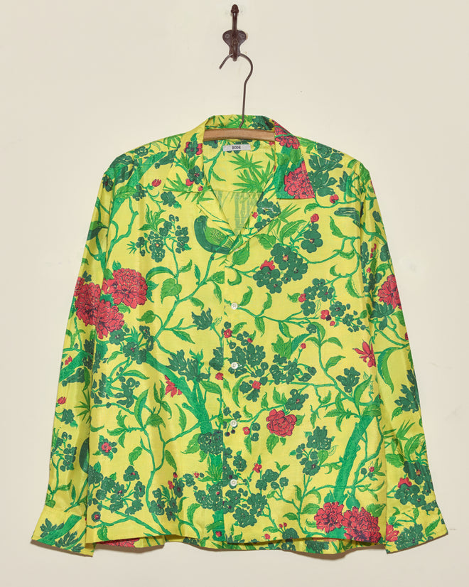The Chartreuse Jungle Shirt (M/L), featuring a unique green and red floral pattern, hangs on a hook against a plain wall.