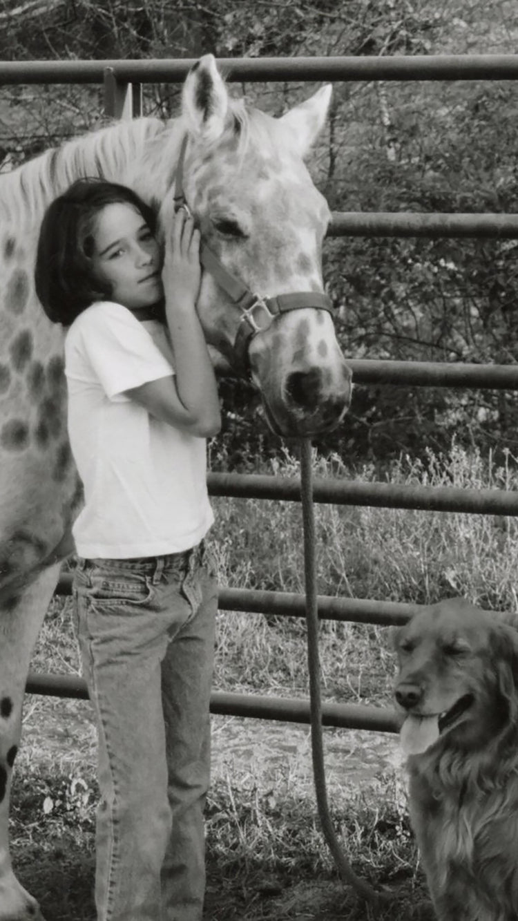 A girl wearing a white tee shirt and jeans hugs her pony while her beloved dog looks on contently.
