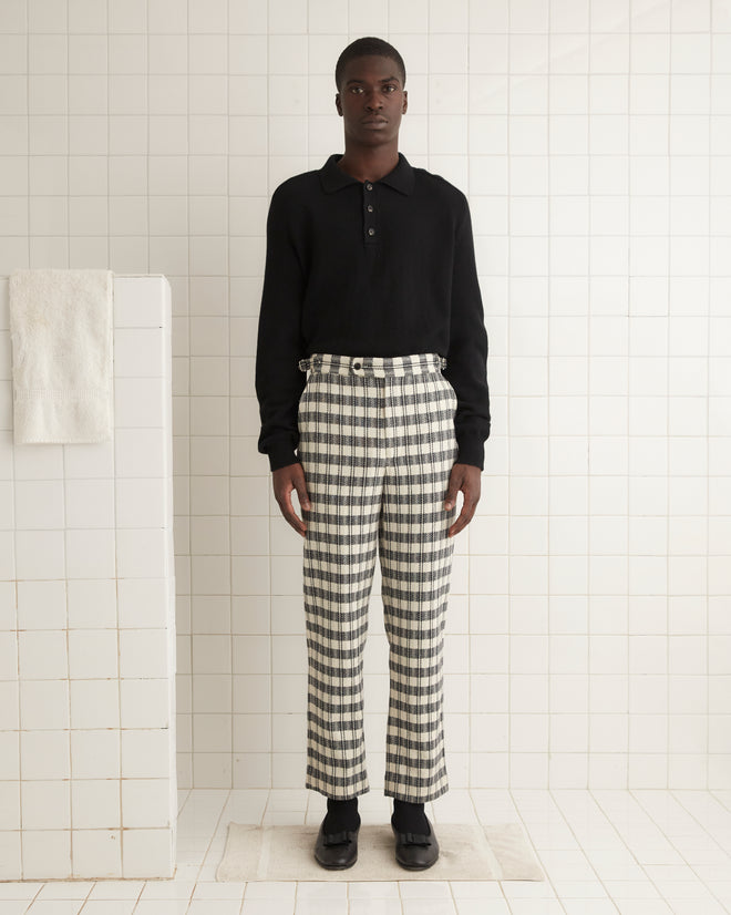 A pair of Check Trousers, made from 100% wool, is folded in half and hung on a wooden hanger against a plain white wall.