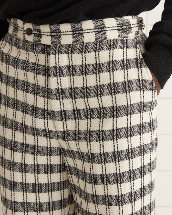 A pair of Check Trousers, made from 100% wool, is folded in half and hung on a wooden hanger against a plain white wall.