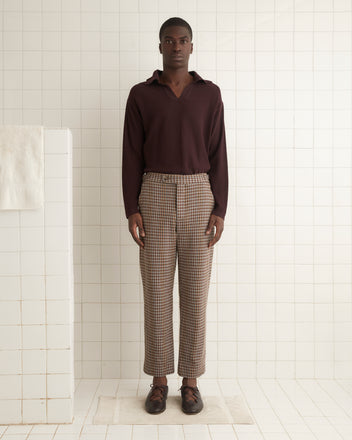 A person with long hair stands against a beige wall, wearing a white button-up shirt, Marston Check Trousers, and brown loafers.