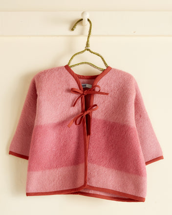 The Chelan Cherry Baby Jacket - OS, featuring pink and red hues with front ties, made in New York, is displayed on a green hanger against a cream wall.