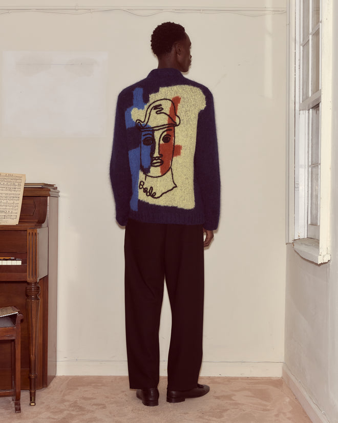 A person in black pants and a superfine Chapeau Cardigan sweater with an abstract face design stands by a piano, facing a wall.