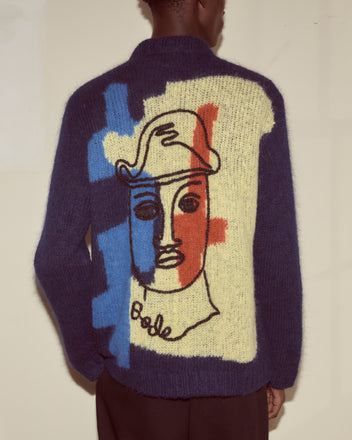 Wearing the Chapeau Cardigan, featuring bold blue, red, and cream colorblocking with an abstract face design on the back—crafted from superfine alpaca for a modernist-inspired, cozy statement piece.