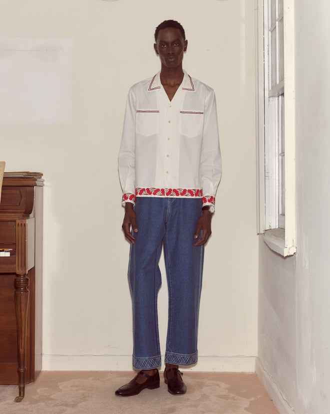 A man stands indoors by a piano and window, wearing the Cherry Lane Beaded Shirt in white paired with blue jeans.