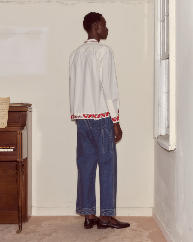 Wearing the Cherry Lane Beaded Shirt, blue jeans, and black shoes—crafted in India—a person stands by a window and an upright piano.