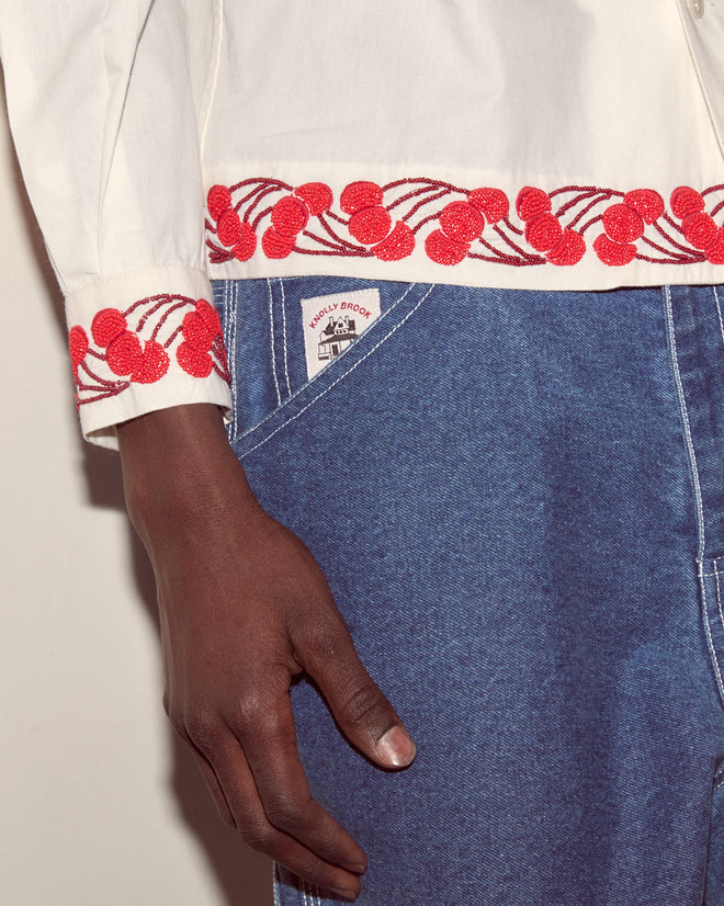 The Cherry Lane Beaded Shirt features cotton poplin cuffs hand-embroidered with beaded cherries, crafted by artisans in India and styled alongside blue jeans.