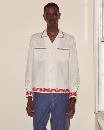 Wearing the Cherry Lane Beaded Shirt in white, a person stands by a beige wall near a window. The shirt features red trim, is made from cotton poplin, paired with blue jeans, and crafted in India.